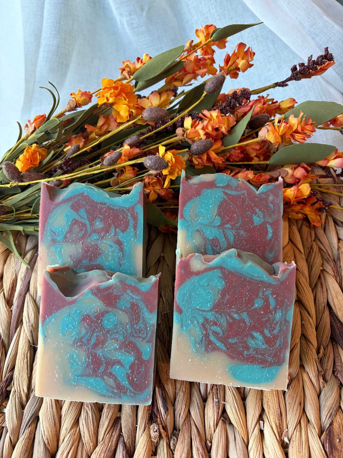 Four handmade marbled soap bars in teal, dusty rose, and cream, arranged on a woven straw mat with orange flowers in the background.