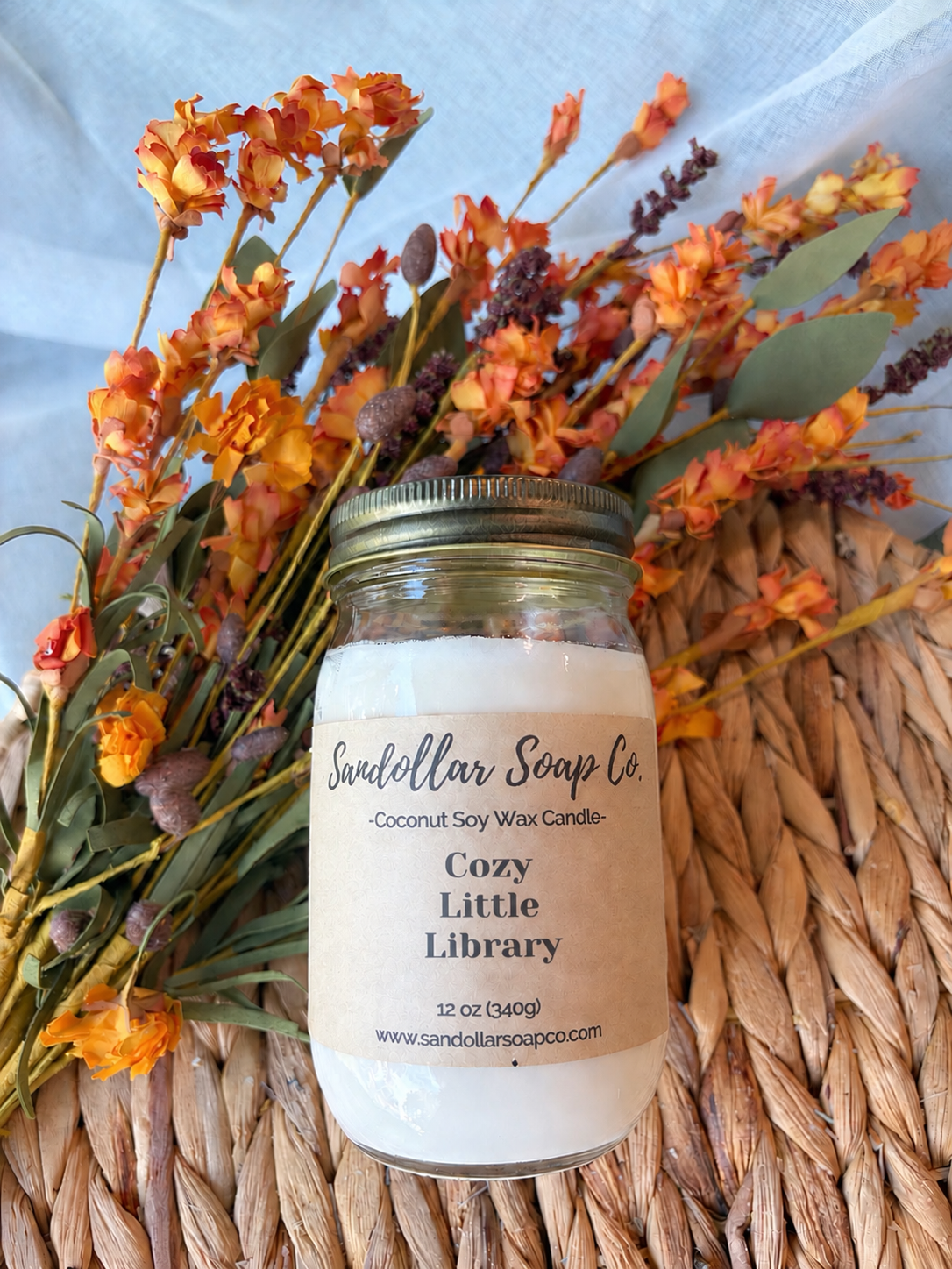 A 12 oz soy wax candle by Sandollar Soap Co. in a clear glass jar with a metal lid, labeled 'Cozy Little Library'.