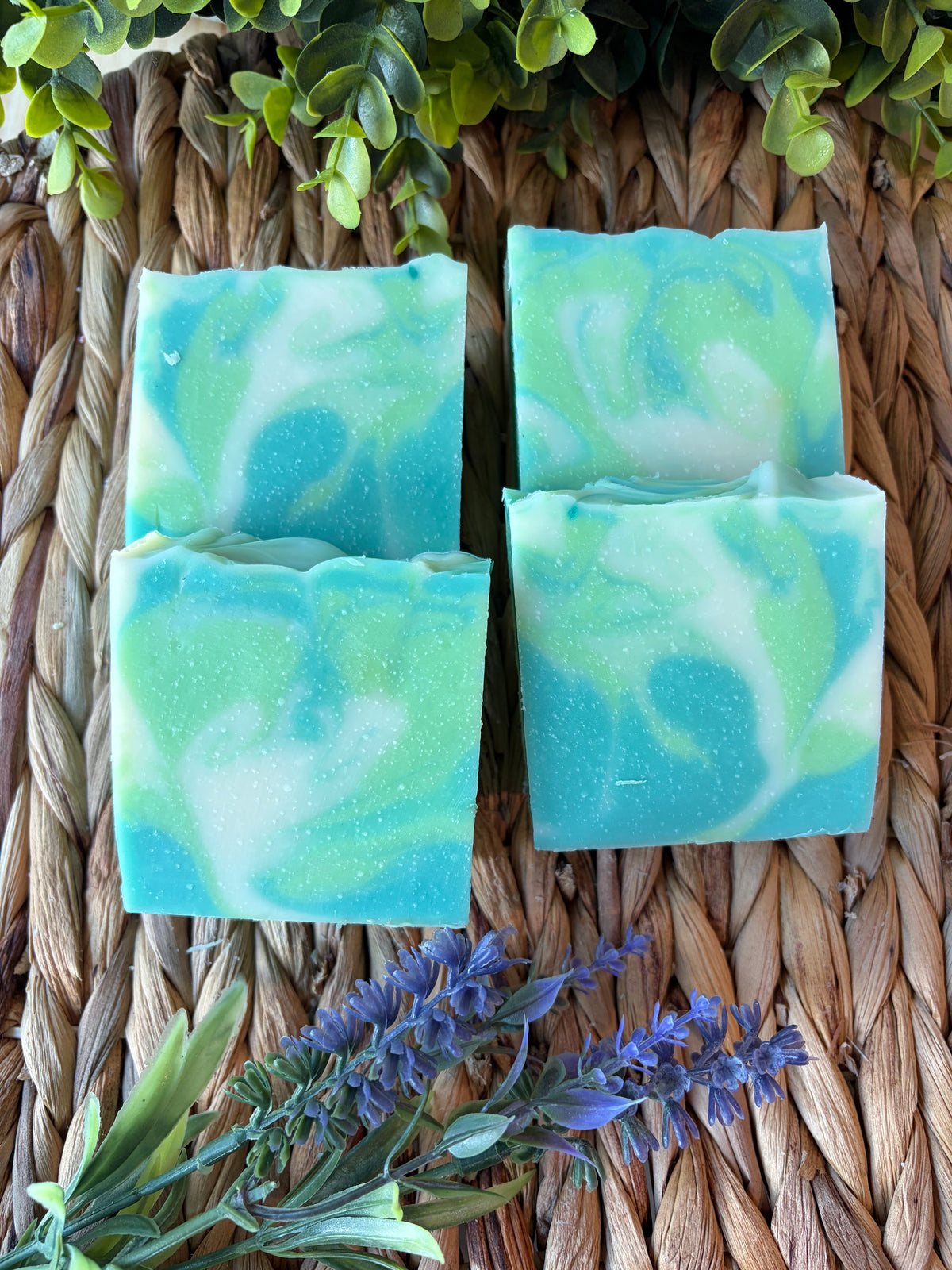 Four rectangular handmade soap bars with a swirled turquoise, mint green, and white pattern, arranged on a woven straw mat with decorative green leaves and purple flowers.
