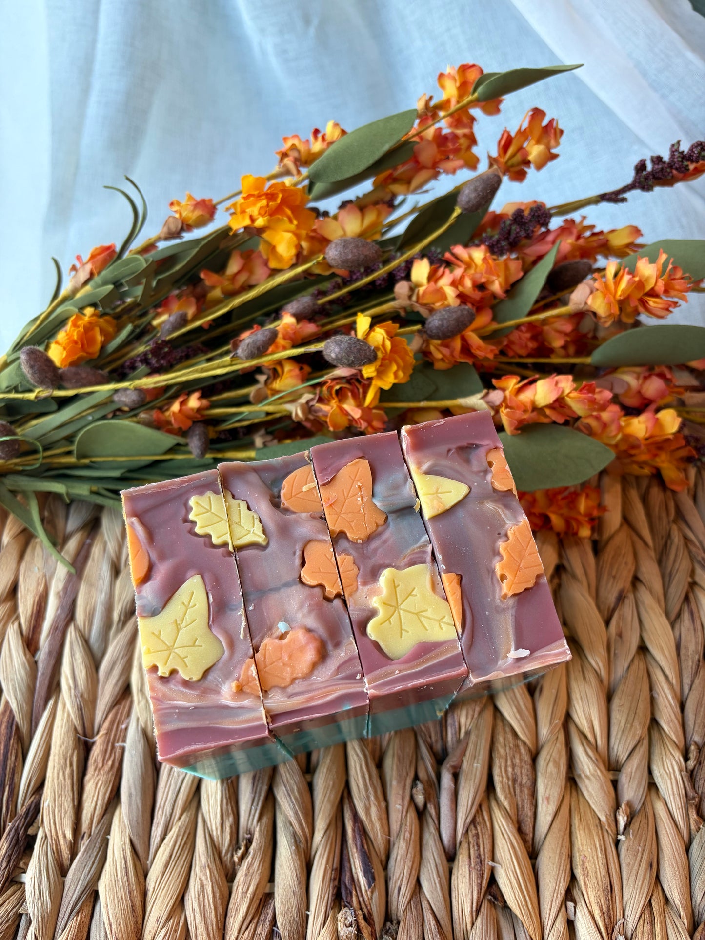Three rectangular bars of handmade soap with embedded orange and yellow autumn leaves, styled on a woven straw mat with dried orange flowers in the background.