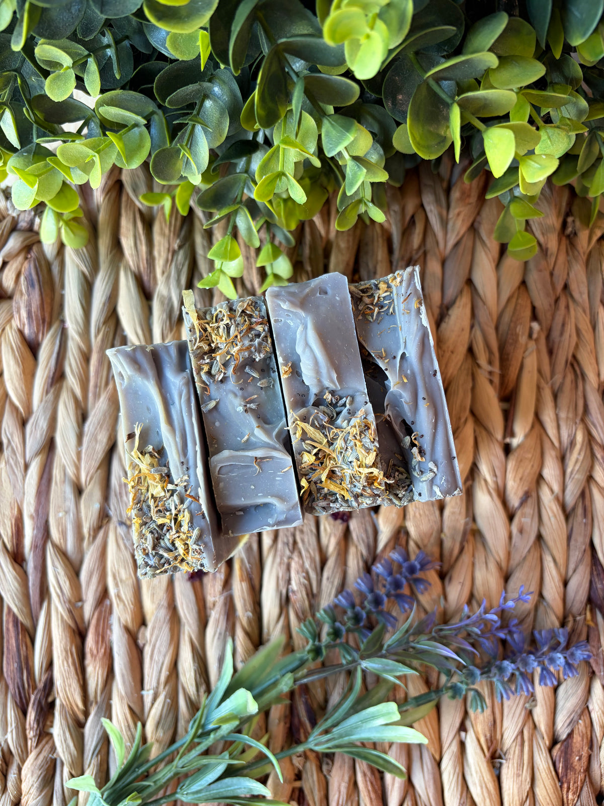 Four rectangular bars of soap with a gray base and dried botanicals embedded along the bottom, arranged on a woven straw mat with green leaves and lavender sprigs around them.