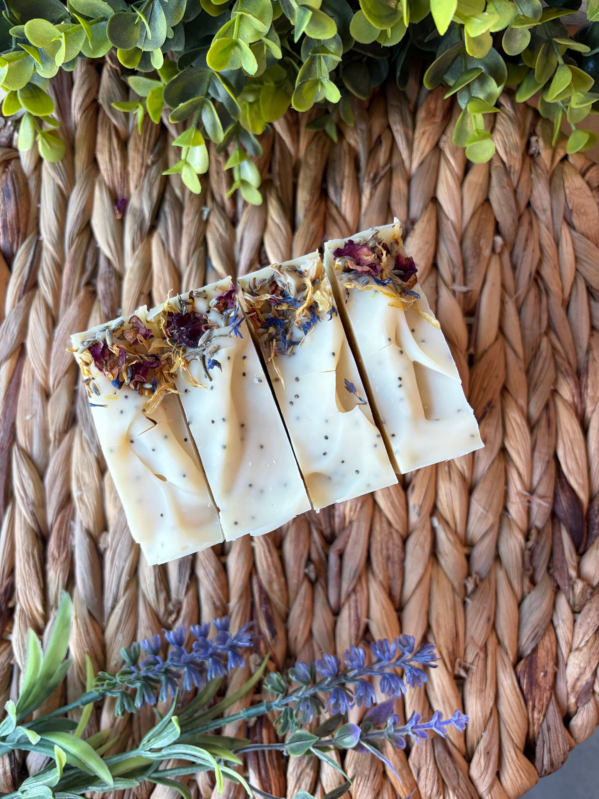 Four rectangular bars of handmade soap with dried flowers and botanicals on top, arranged on a woven straw mat with green leaves and lavender sprigs.