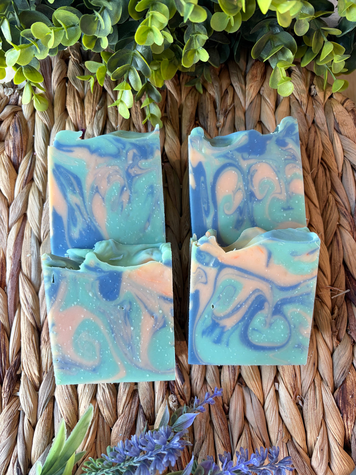 Two handmade marbled soap bars in shades of blue, teal, and peach, arranged on a woven straw mat with green leaves and purple flowers as decorative props.