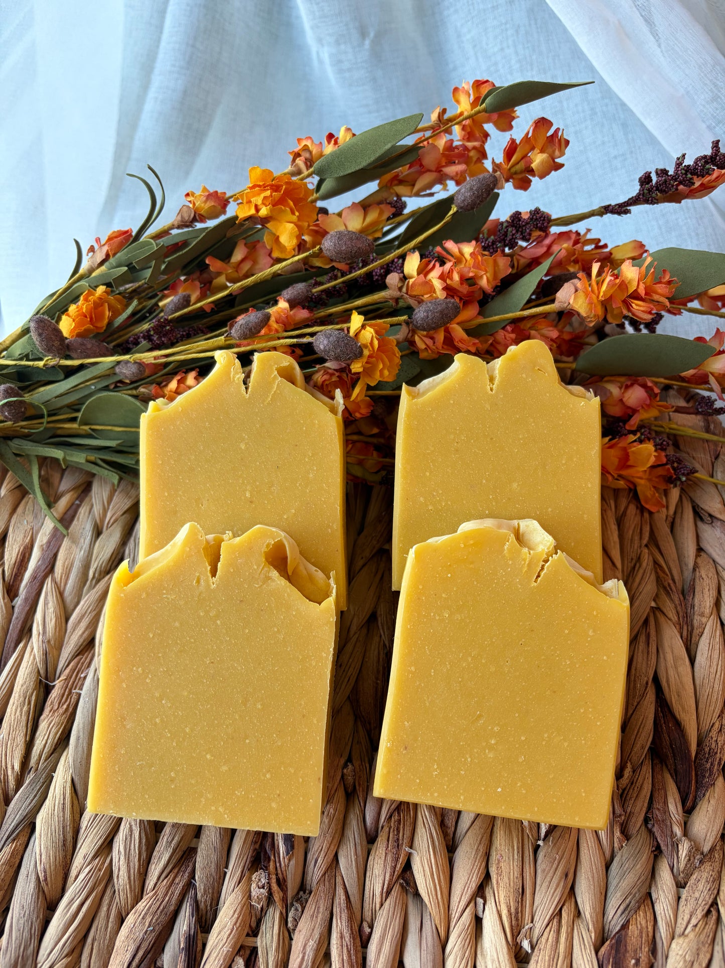 Four rectangular bars of handmade yellow soap with rustic, jagged tops, arranged on a woven straw mat with dried orange flowers in the background.