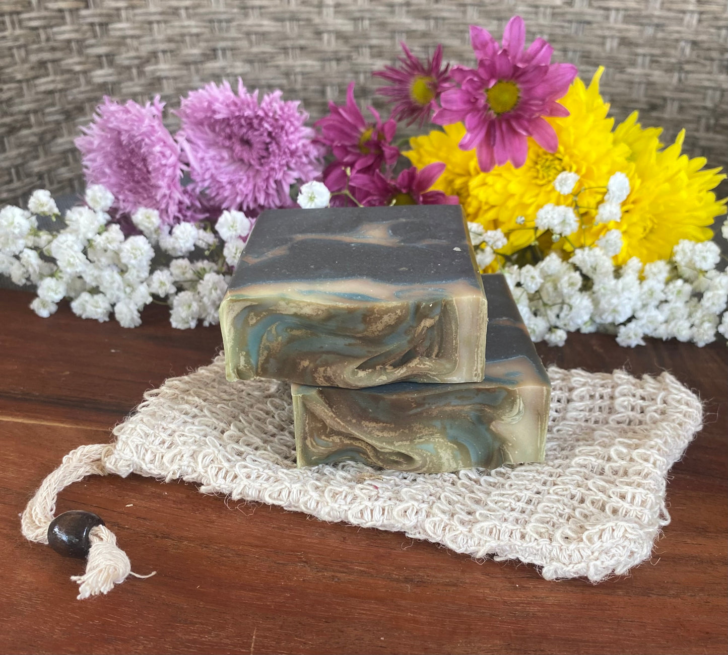 Handmade bar soap with a swirled blue and white design, placed on a beige loofah on a wooden surface, with colorful flowers in the background.