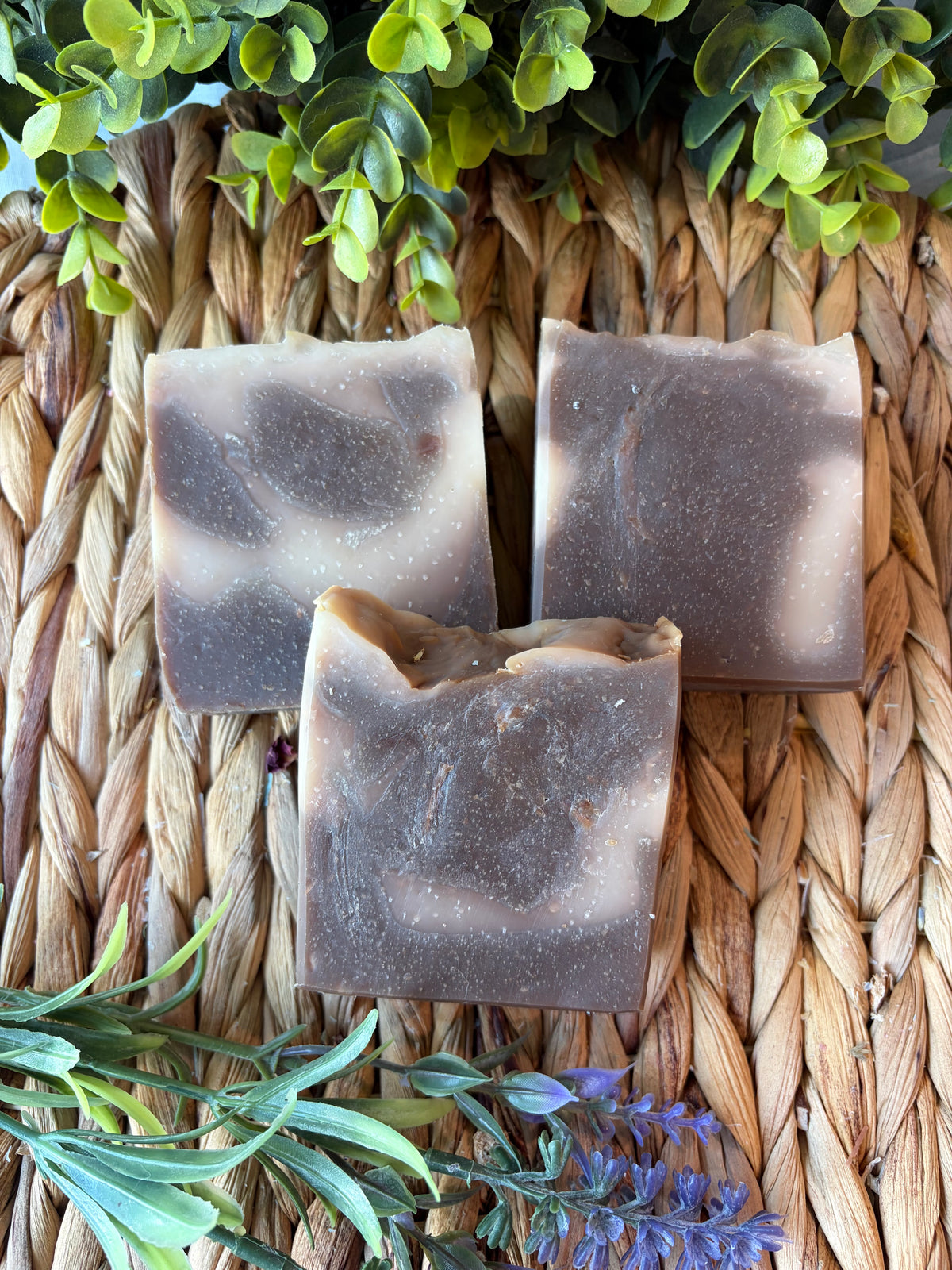 Three handmade rectangular soap bars with a brown and beige marbled pattern, arranged on a woven straw mat with green and purple botanical accents.