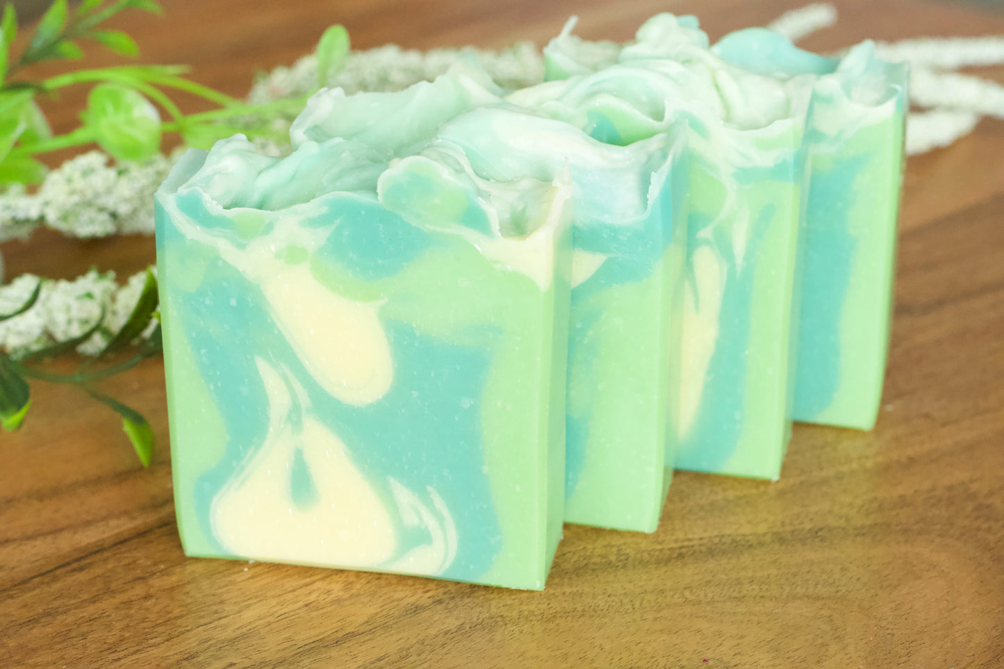 Hand-cut bars of cucumber and green tea soap with a white and green marbled design, displayed on a wooden surface with a sprig of flowers in the background.