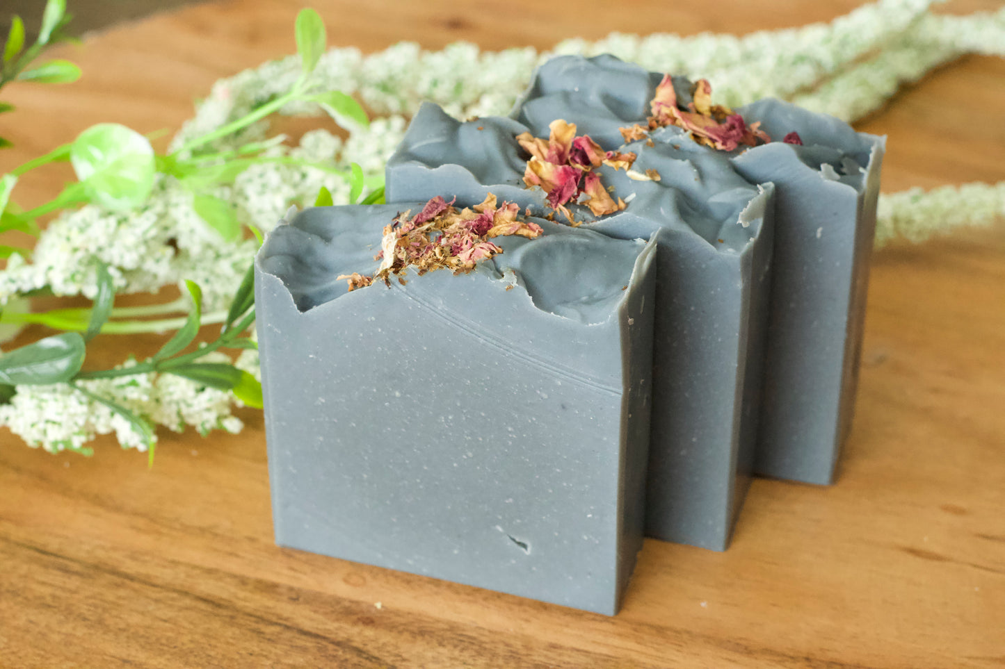Hand-cut bars of activated charcoal facial soap with dried rose petals on top, displayed on a wooden surface with a white flower in the background.