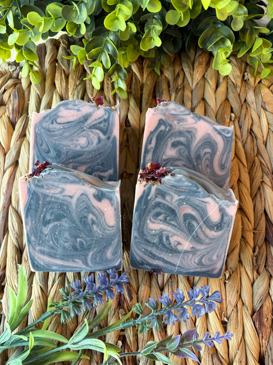 Four rectangular artisan soap bars with a swirled gray and pale pink pattern and small dried flower accents, arranged on a woven straw mat with green leaves and lavender sprigs around them.