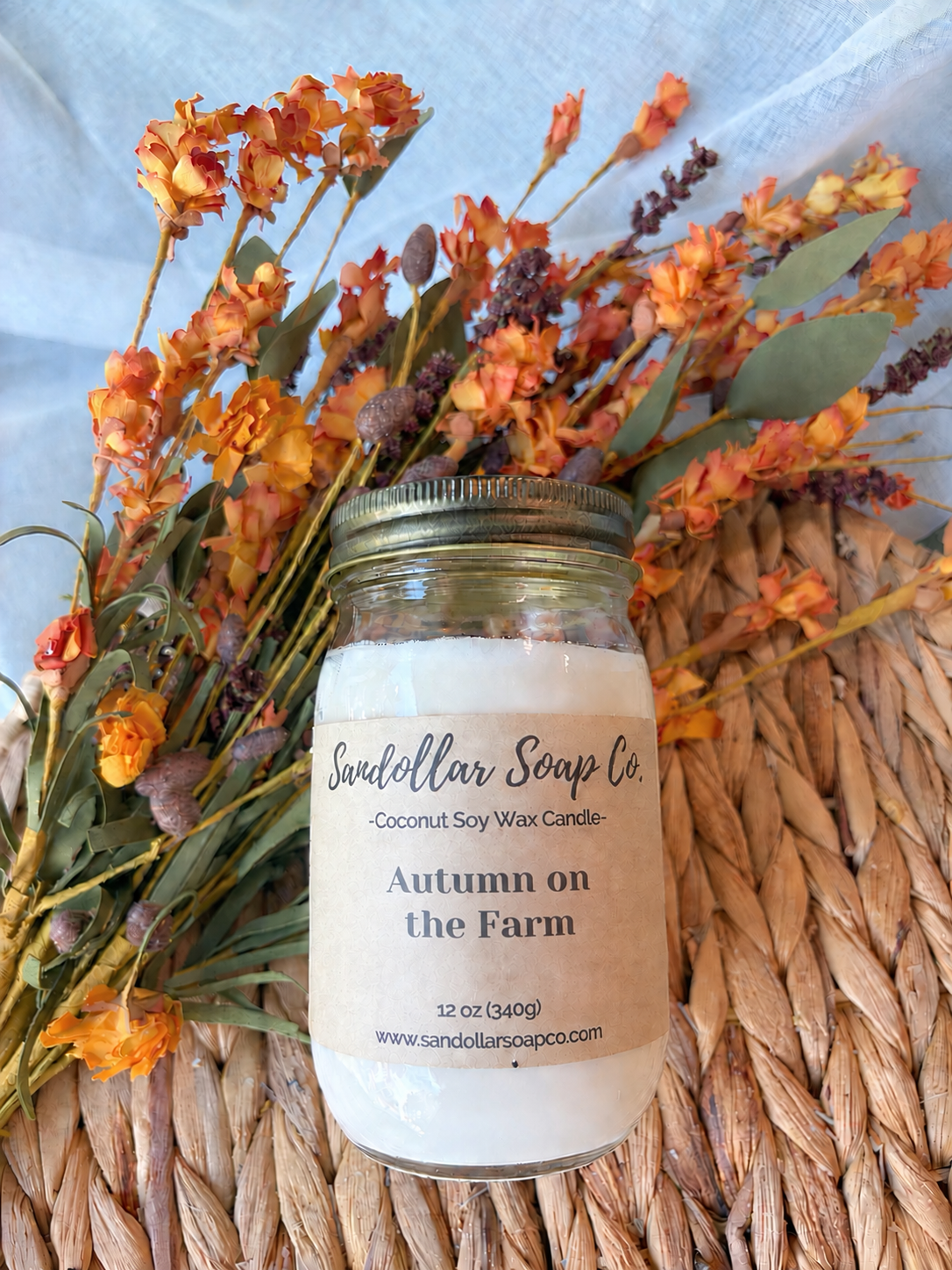 A jar candle labeled 'Autumn on the Farm' by Sandollar Soap Co., placed on a woven straw mat with dried orange and yellow flowers behind it against a light blue fabric backdrop.