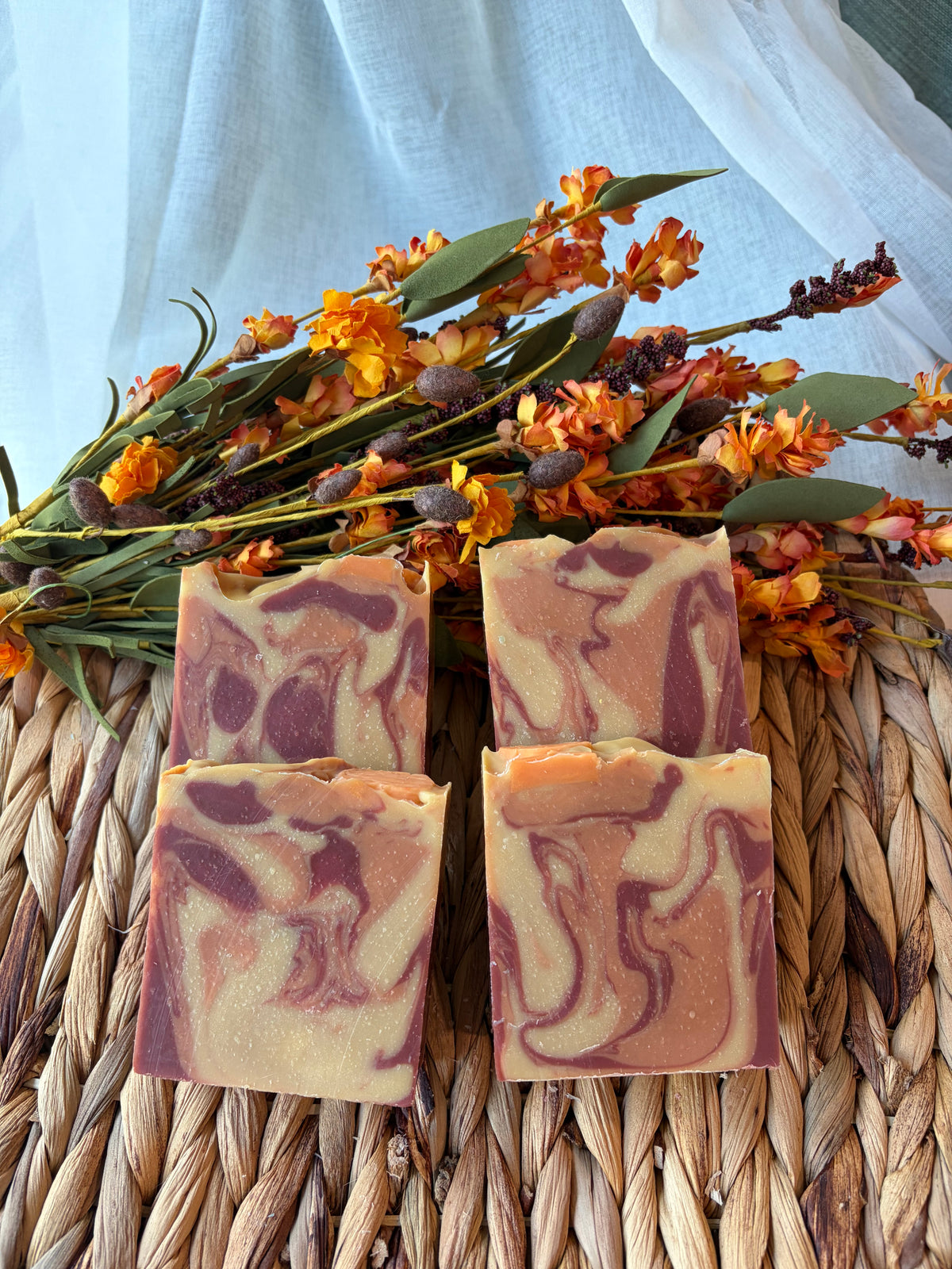 Four rectangular bars of marbled handmade soap in peach, cream, and plum tones, arranged on a woven straw mat with orange flowers and greenery behind them.