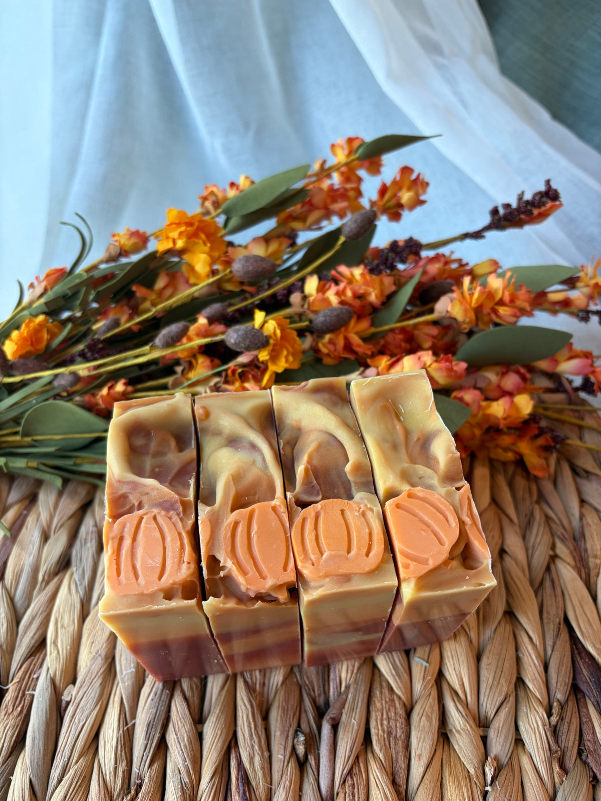 Four artisanal pumpkin-shaped soaps with orange and cream marbled swirls, arranged on a woven straw mat with dried orange flowers in the background.
