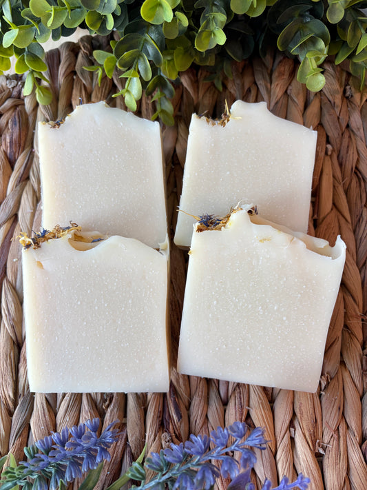 Four rectangular bars of handmade soap with rustic broken edges, decorated with dried botanicals, arranged on a woven brown basket with green leaves and purple flowers.