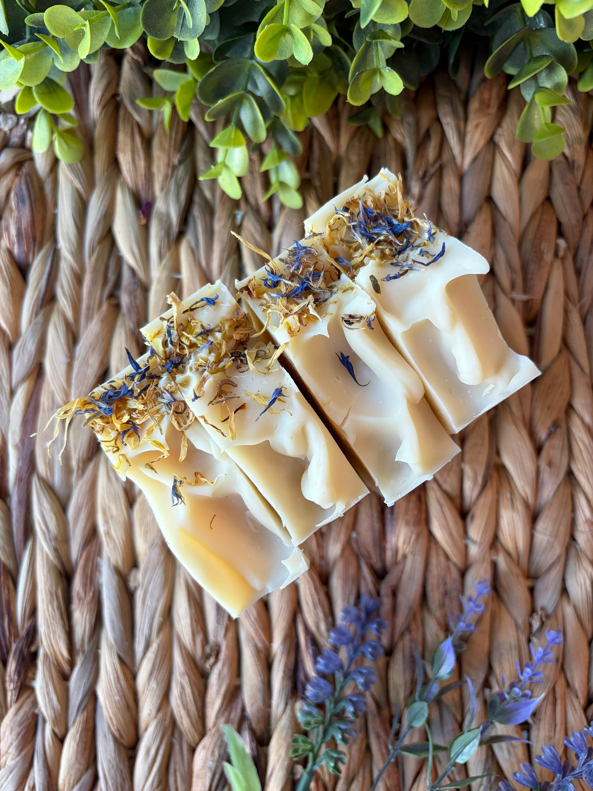 Three rectangular bars of handmade soap with a creamy off-white base, topped with dried yellow botanicals and blue flower petals, arranged on a woven straw mat with green leaves and purple flowers.