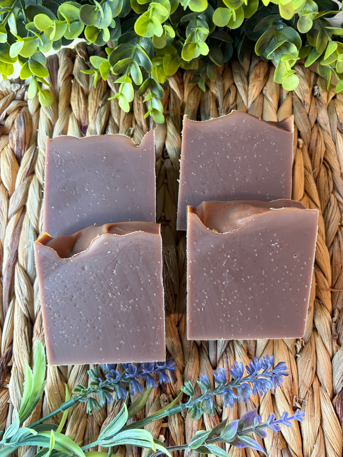 Four rectangular bars of handmade soap in a muted mauve-brown color, arranged on a woven straw mat with green leaves and purple flowers as decorative props.