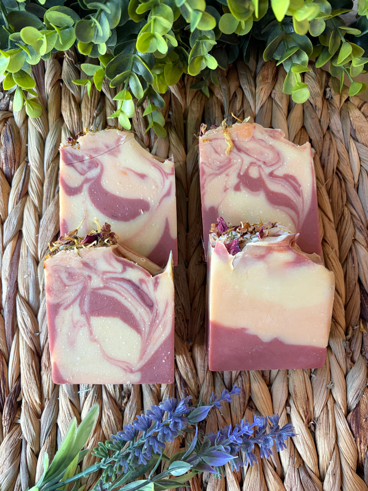 Four artisanal marbled soap bars with dried flower petals on top, arranged on a woven straw mat with green leaves and purple flowers as decorative props.
