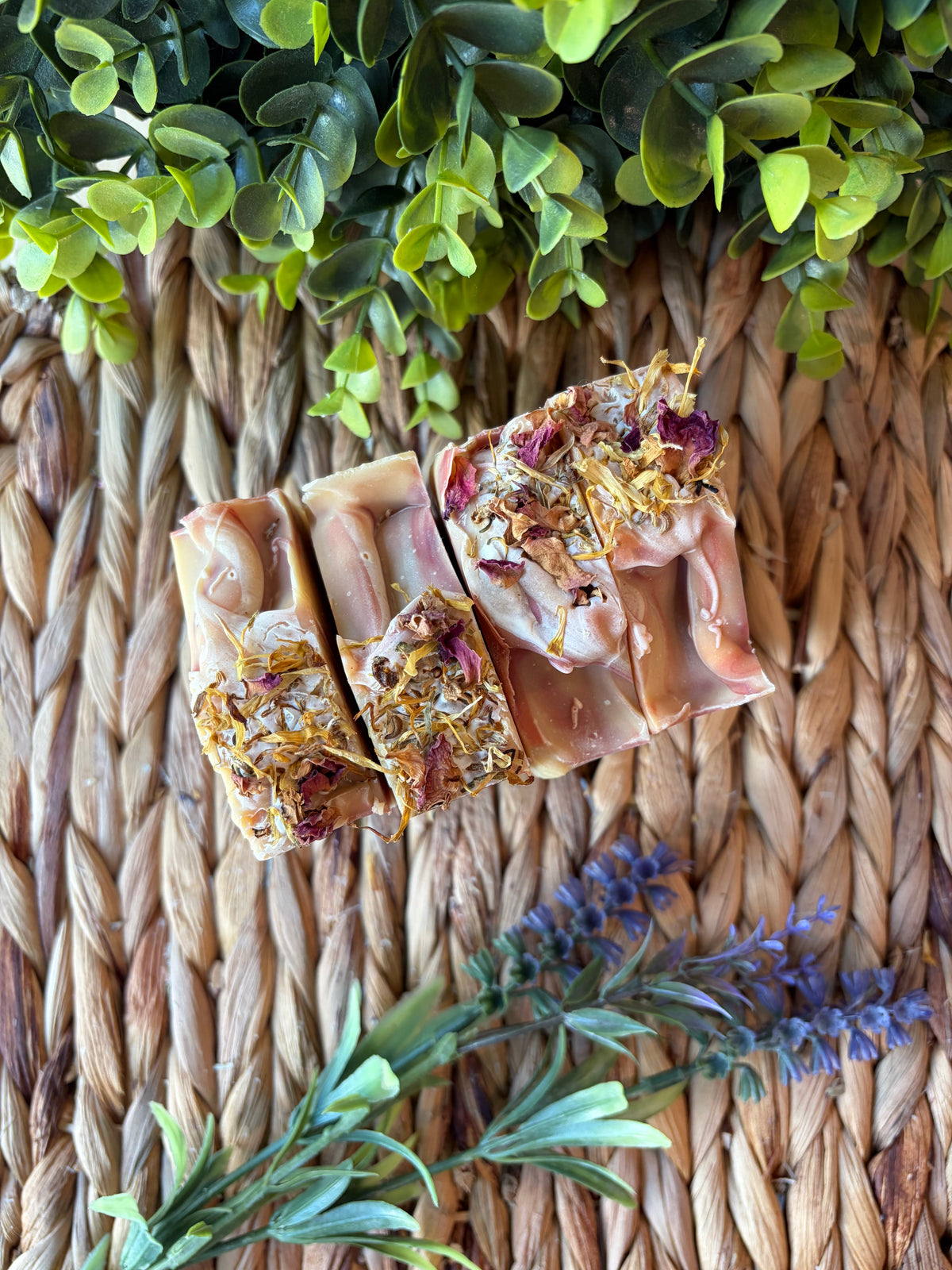 Three rectangular bars of handmade soap with dried flower and herb inclusions, arranged on a woven straw mat with green leaves and purple flowers as decorative props.