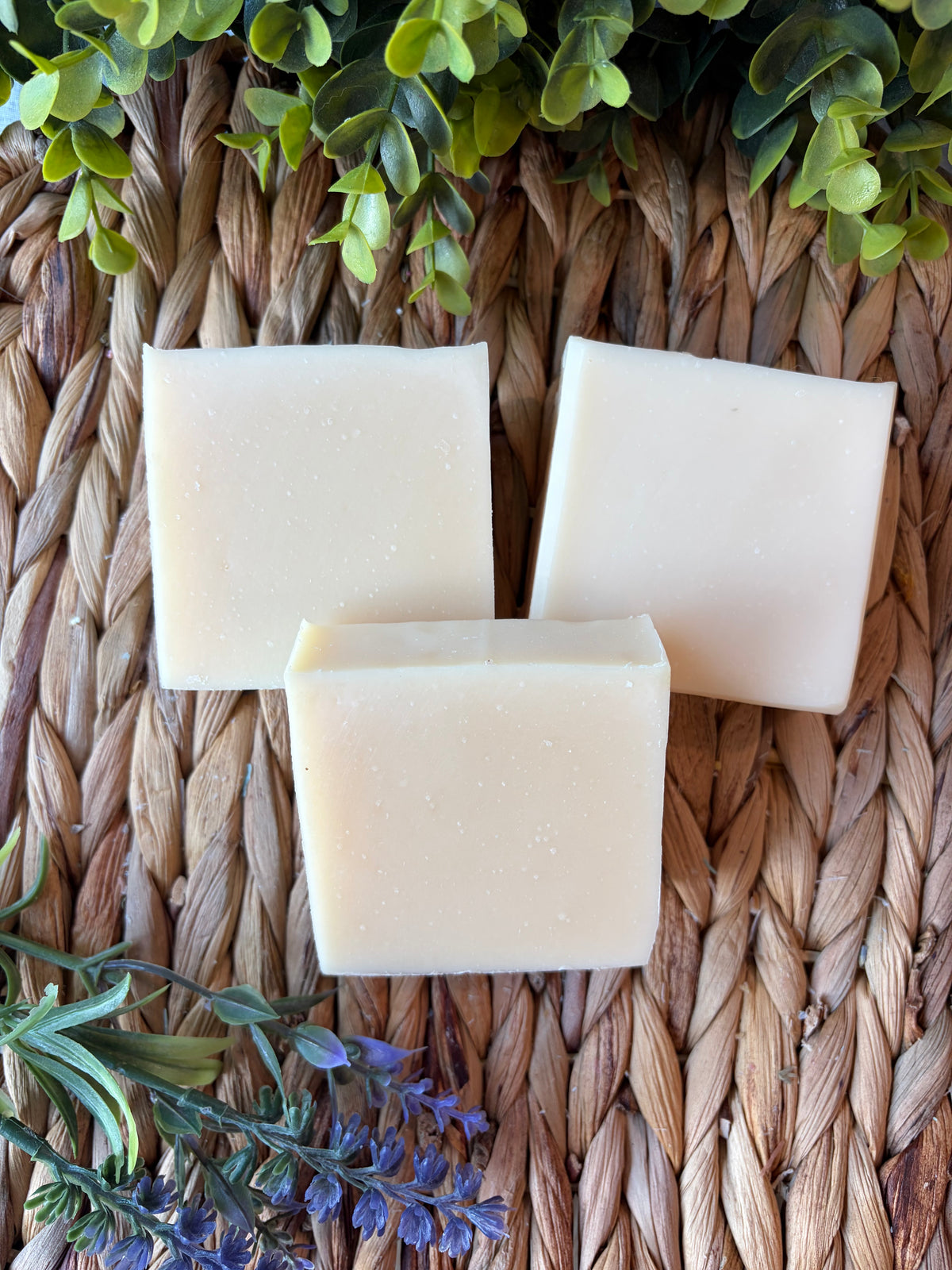 Three rectangular bars of pale cream-colored soap arranged on a woven straw mat, with green leaves at the top and small purple flowers at the bottom.