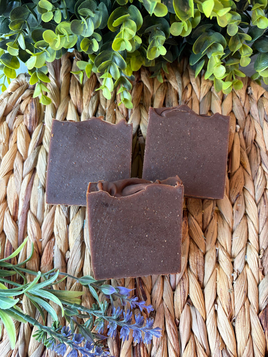 Three rectangular bars of dark brown handmade soap arranged on a woven straw mat, with green foliage and small purple flowers used as styled props.