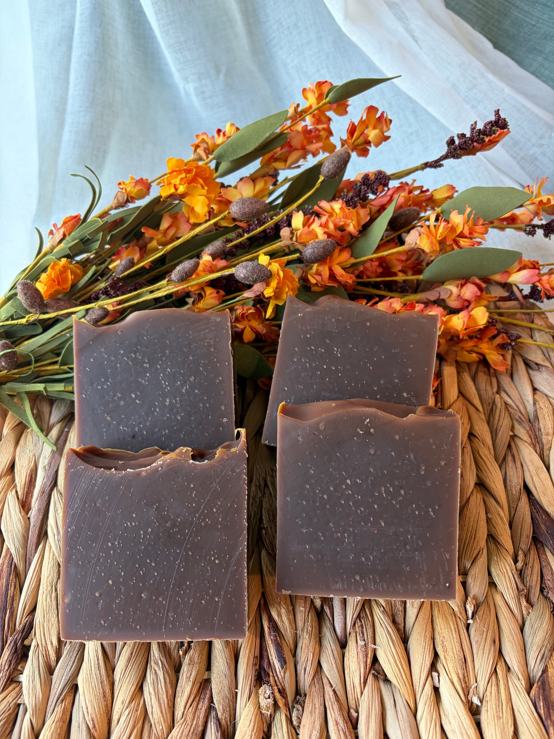Four rectangular bars of dark brown handmade soap with a speckled texture, arranged on a woven straw mat with dried orange flowers and green leaves in the background.