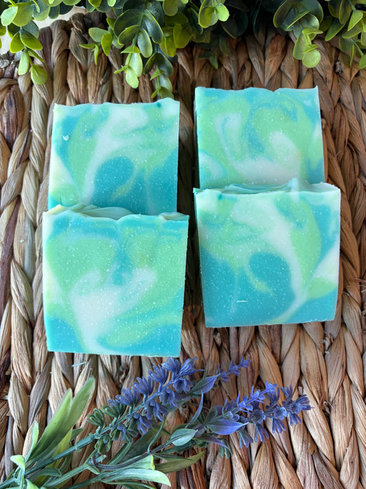 Four rectangular handmade soap bars with a swirled turquoise, mint green, and white pattern, arranged on a woven straw mat with decorative green leaves and purple flowers.