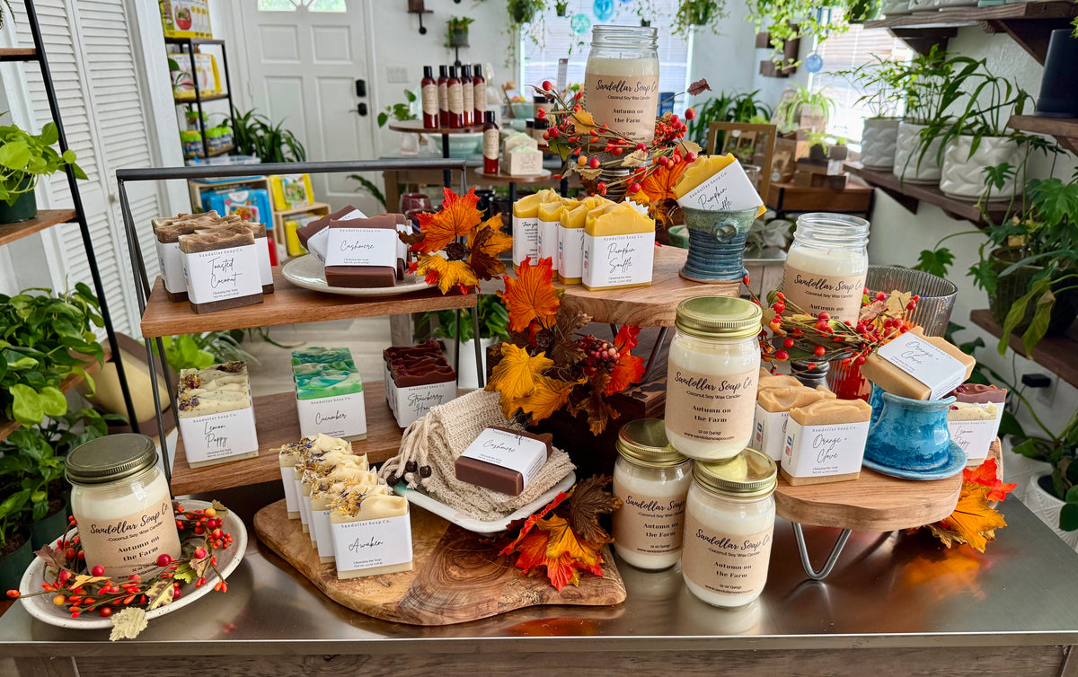 A styled product display of handmade soaps and candles on a metal counter inside a shop. Multiple cream-colored candles in glass jars and bars of soap in boxes and wrapped pieces are arranged on wooden stands and trays, decorated with autumn leaves and small floral accents.