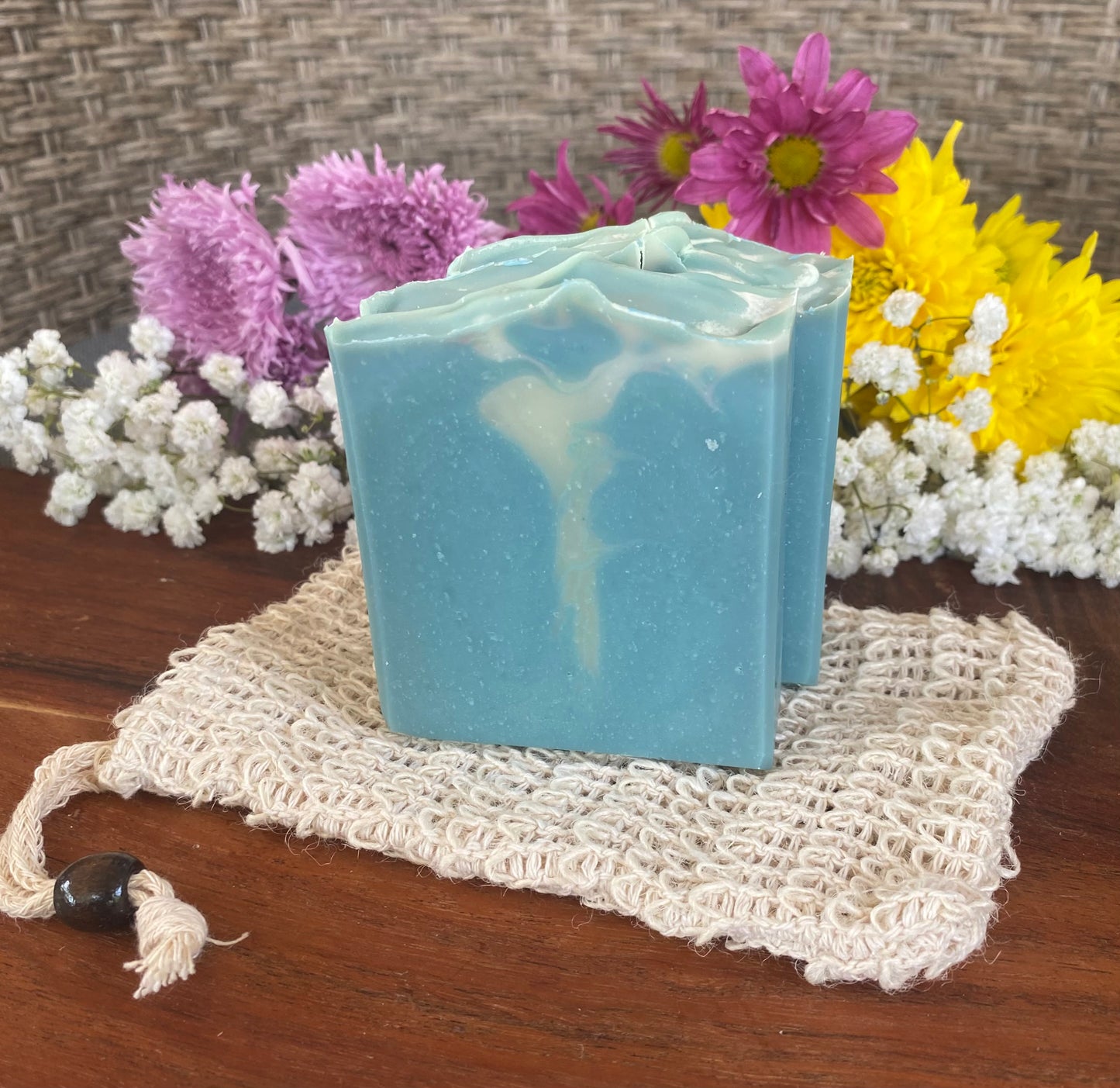 A bar of soap with a turquoise hue placed on a beige, square-shaped sponge, with a background that includes colorful flowers and a wooden surface.