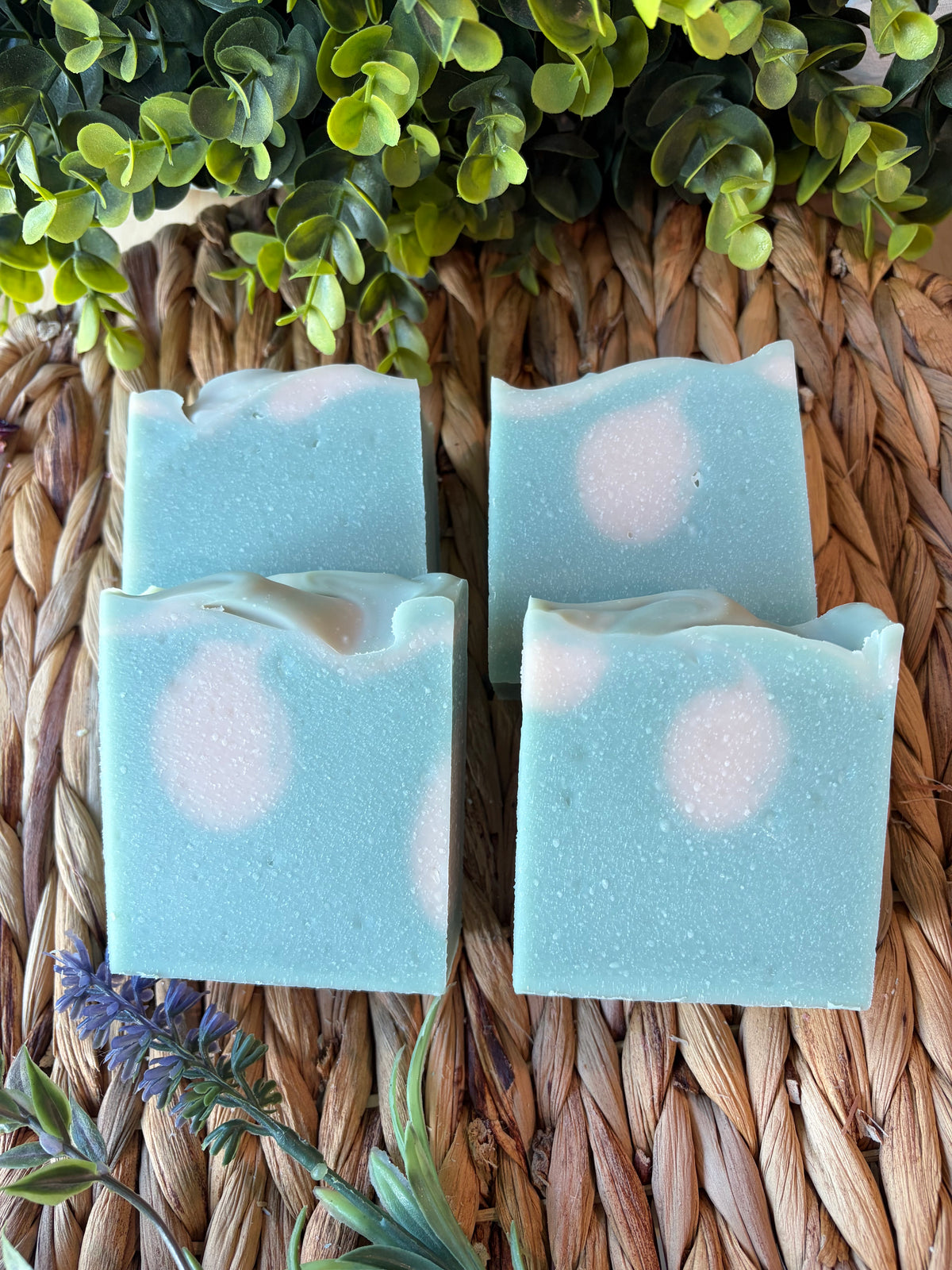 Four rectangular bars of handmade soap in a light blue color with white circular swirls, arranged on a woven straw mat with green foliage and lavender sprigs.