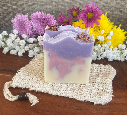 A bar of soap with a purple top layer, Pink middle layer, and a beige bottom layer, garnished with dried rose petals, resting on a soap saver with flowers in the background.