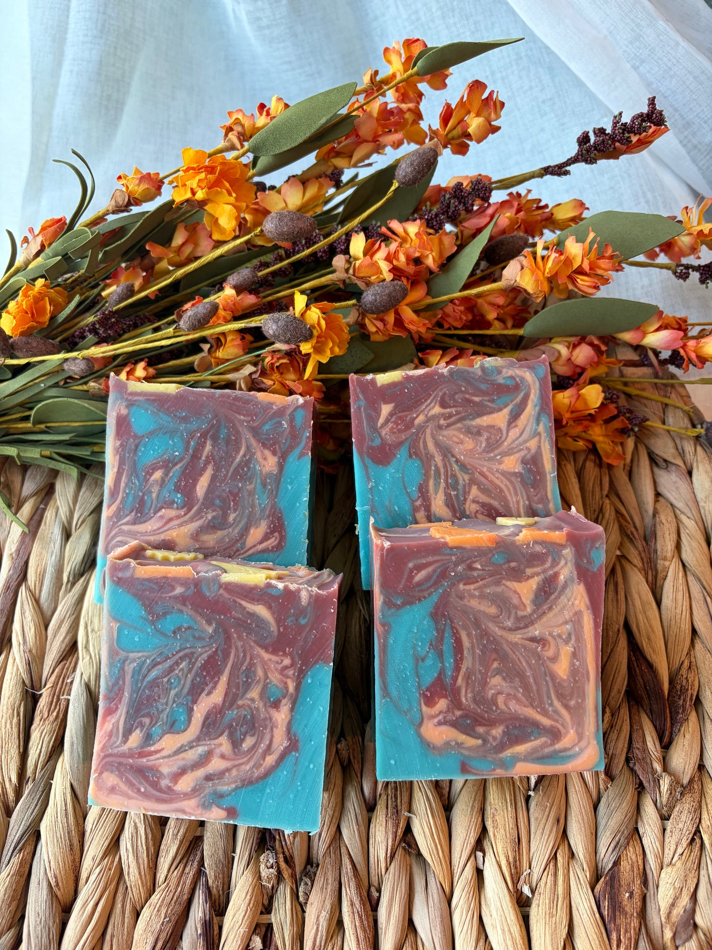 Four rectangular handmade soap bars with a marbled swirl of turquoise blue, terracotta red, and peach-orange, arranged on a woven straw mat with dried orange flowers in the background.