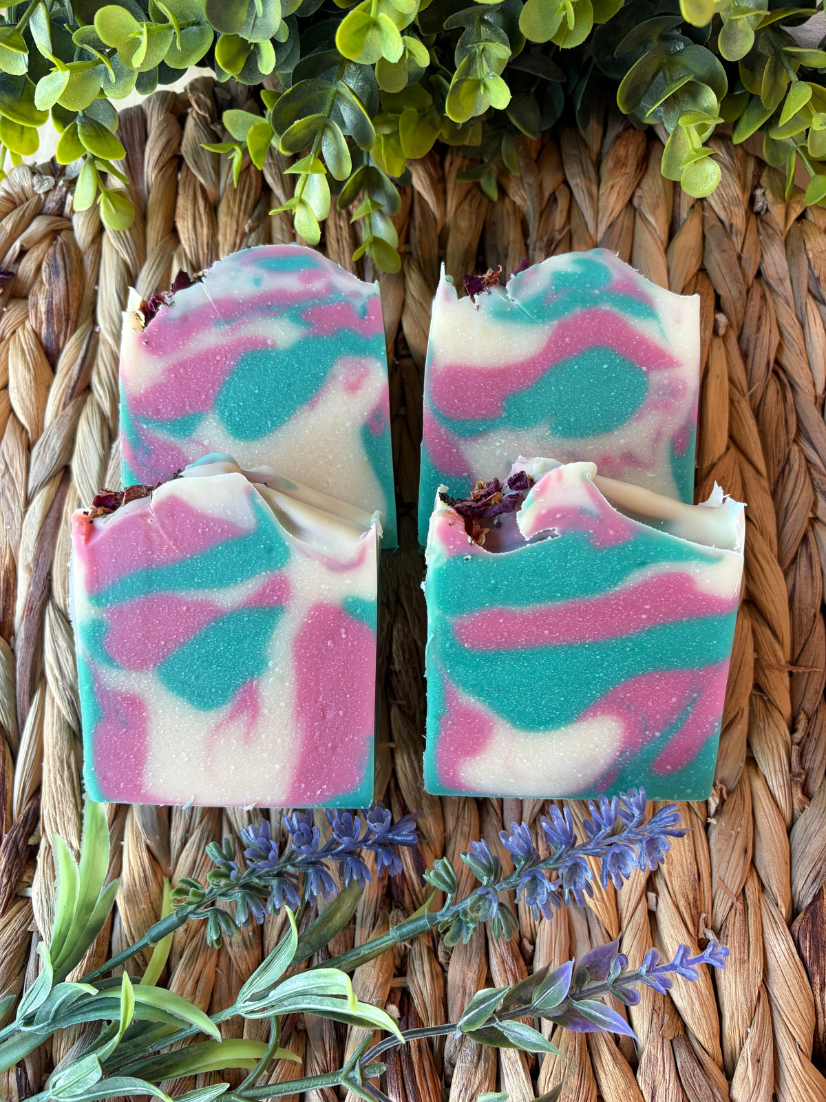 Four marbled artisan soap bars with pink, teal, and white swirls, arranged on a woven straw mat with green leaves and lavender sprigs.