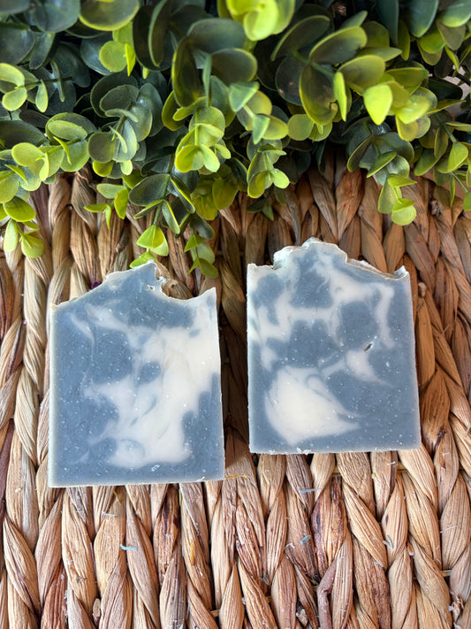 Two handmade rectangular bars of soap with a gray and white marbled pattern, displayed on a woven straw mat with green foliage at the top.