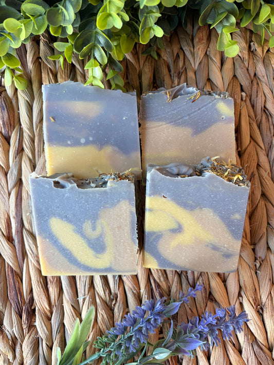 Four rectangular artisan soap bars with a marbled gray and yellow pattern, displayed on a woven straw mat with green leaves and lavender sprigs.