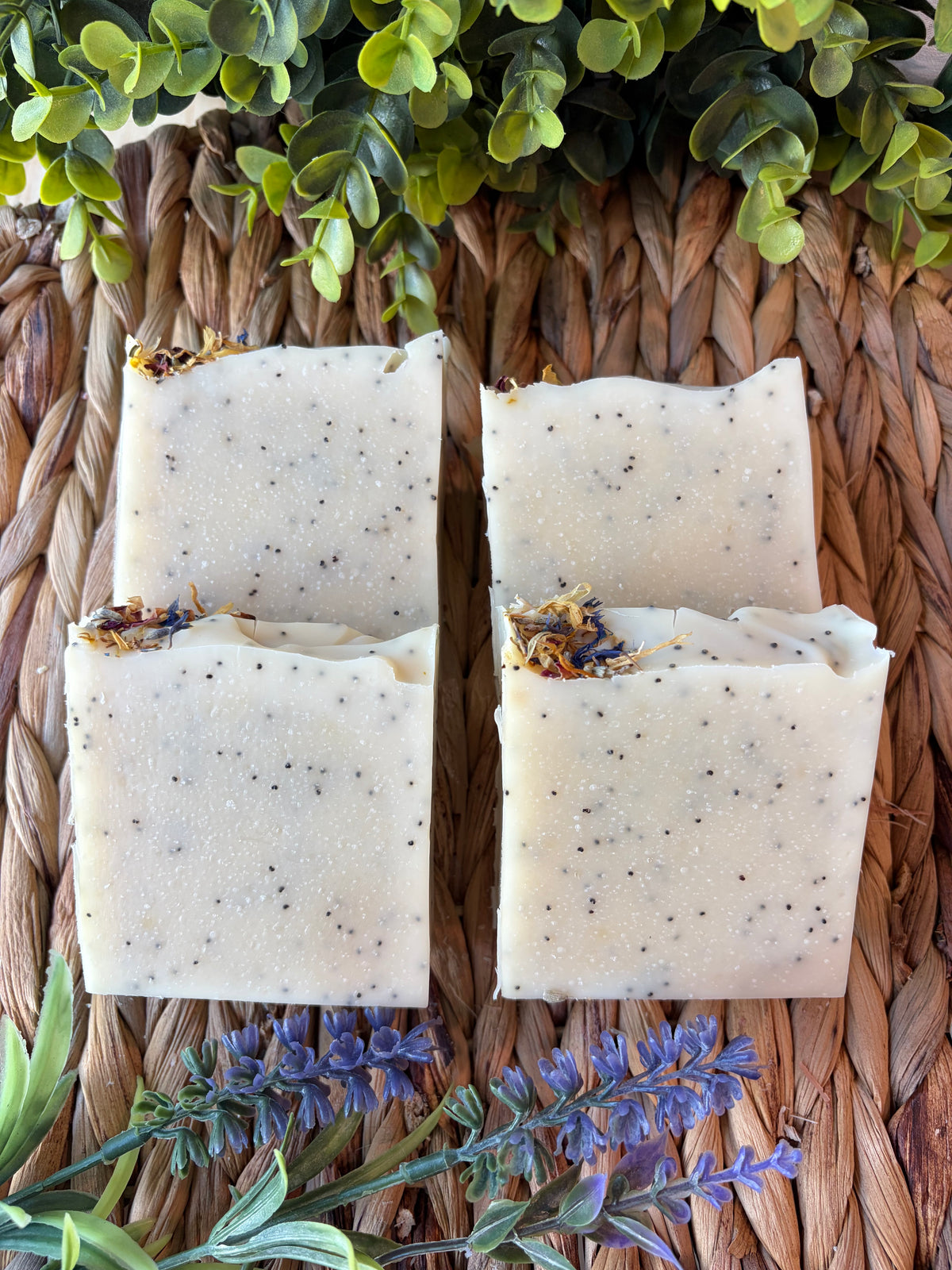 Four rectangular bars of handmade soap with speckled dark flecks and dried botanical inclusions, arranged on a woven brown mat with green foliage and purple flowers around them.