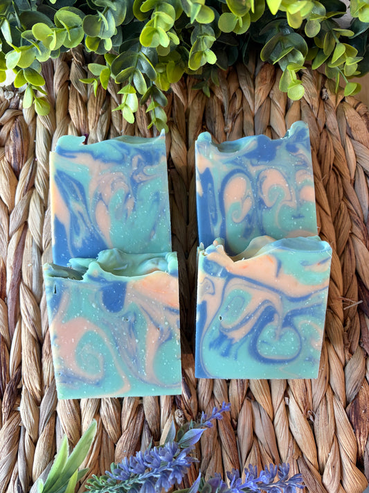 Two handmade marbled soap bars in shades of blue, teal, and peach, arranged on a woven straw mat with green leaves and purple flowers as decorative props.