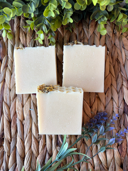 Three rectangular bars of handmade soap in a warm cream-beige color, each topped with a small piece of dried flower, arranged on a woven straw mat with green leaves and purple flowers as decorative props.