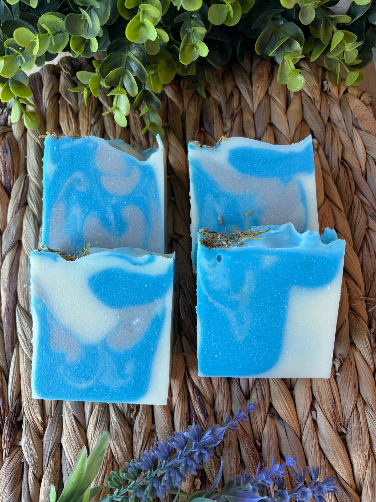 Four handmade rectangular bars of blue and white marbled soap arranged on a woven straw mat, with green leaves and purple flowers used as decorative props.