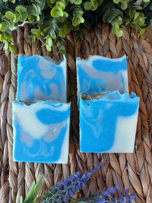Four handmade rectangular bars of blue and white marbled soap arranged on a woven straw mat, with green leaves and purple flowers used as decorative props.