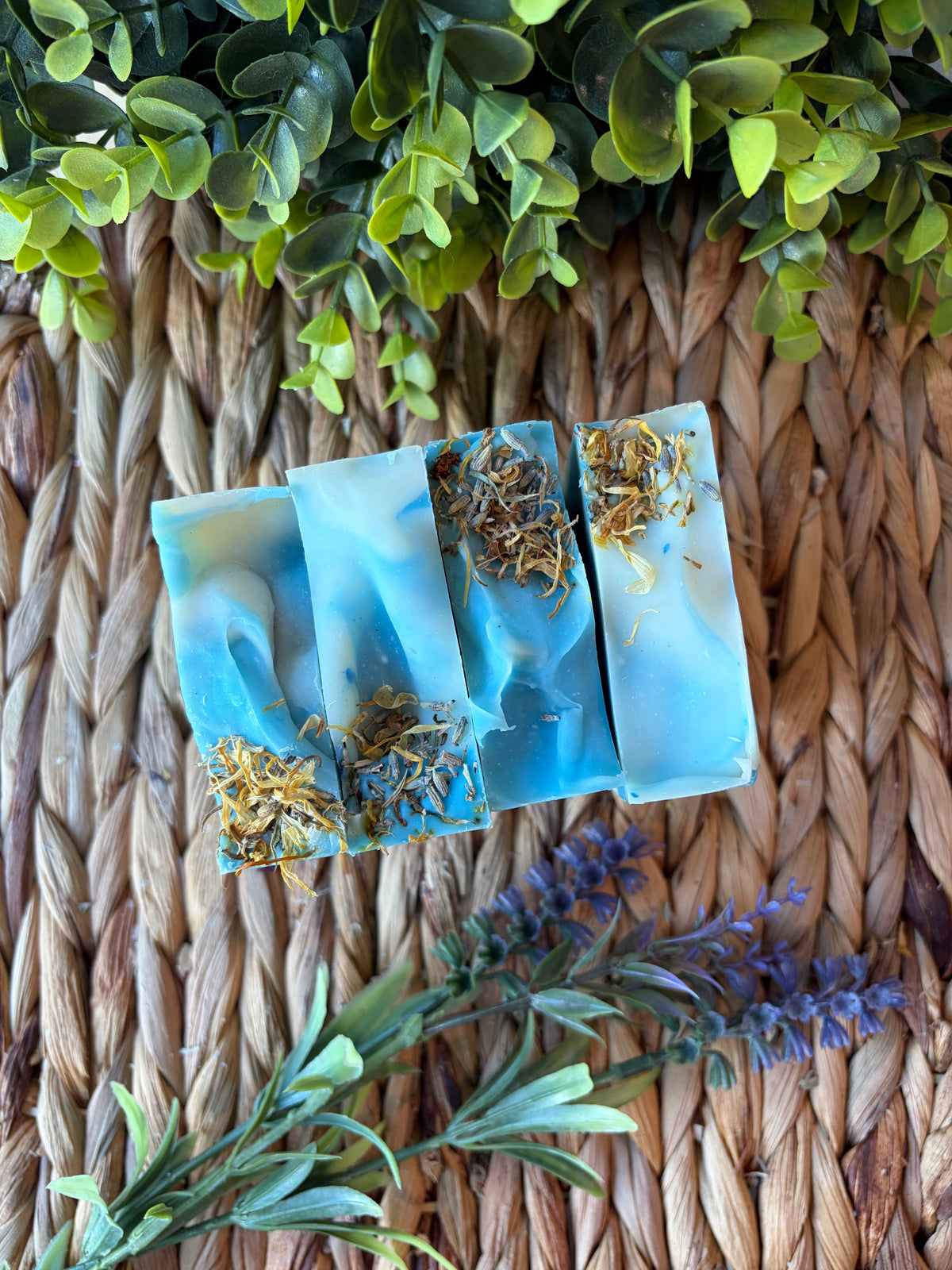 Four rectangular bars of handmade blue soap with dried yellow and orange botanical inclusions, arranged on a woven straw mat with green leaves and purple flowers around them.