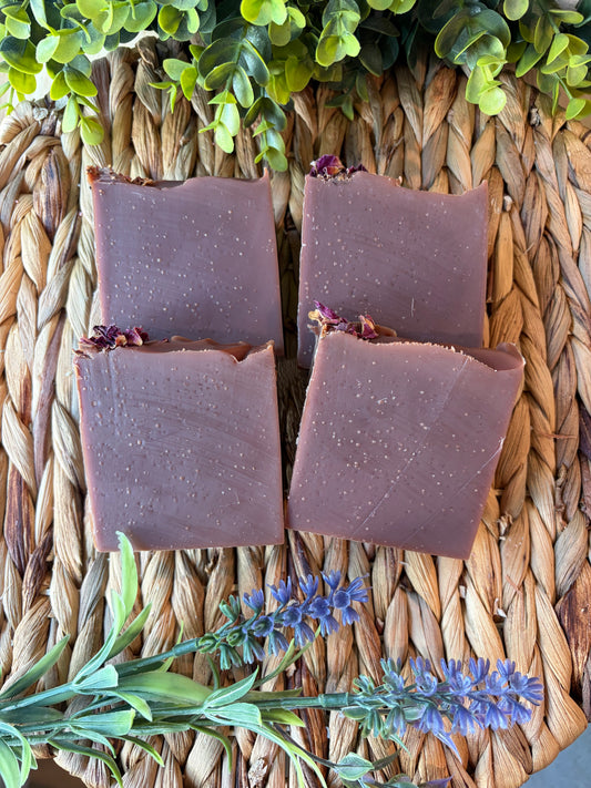Four rectangular bars of handmade soap in a dusty mauve-purple color, arranged on a woven straw mat with green leaves and purple flowers as decorative props.