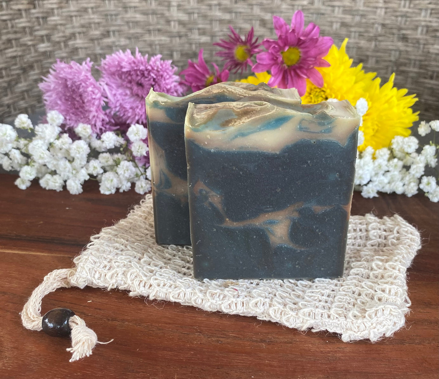 Handmade bar soap with a swirled blue and white design, placed on a beige loofah on a wooden surface, with colorful flowers in the background.