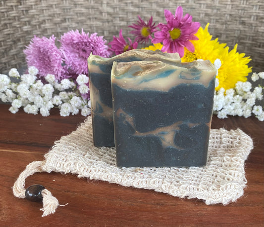Handmade bar soap with a swirled blue and white design, placed on a beige loofah on a wooden surface, with colorful flowers in the background.