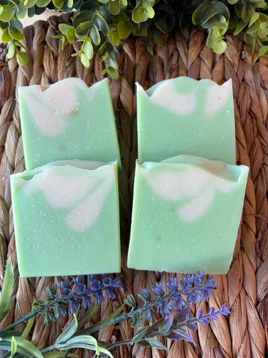 Four rectangular bars of handmade soap in a light mint green color with white marbled swirls, arranged on a brown woven mat with decorative green leaves and purple flowers.