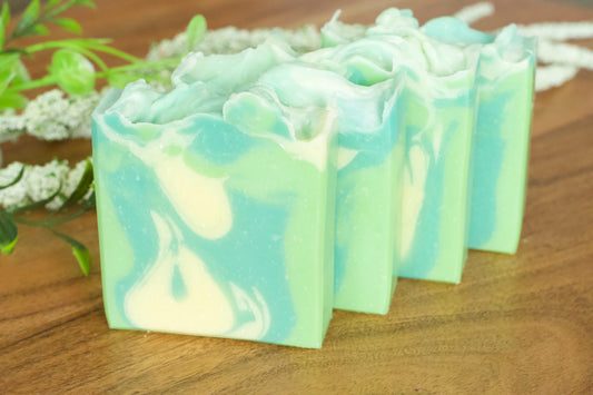 Hand-cut bars of cucumber and green tea soap with a white and green marbled design, displayed on a wooden surface with a sprig of flowers in the background.