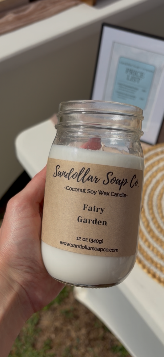 Hand holding a candle labeled Fairy Garden with a blurred background.