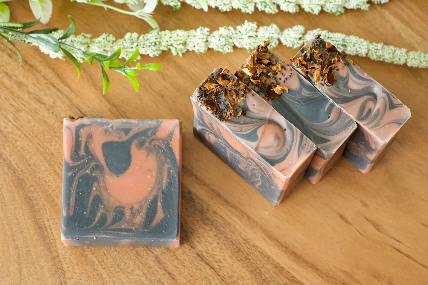 Hand-cut bars of soap with a swirled pattern design, predominantly in shades of gray and pink, displayed on a wooden surface with a white floral decoration in the background.