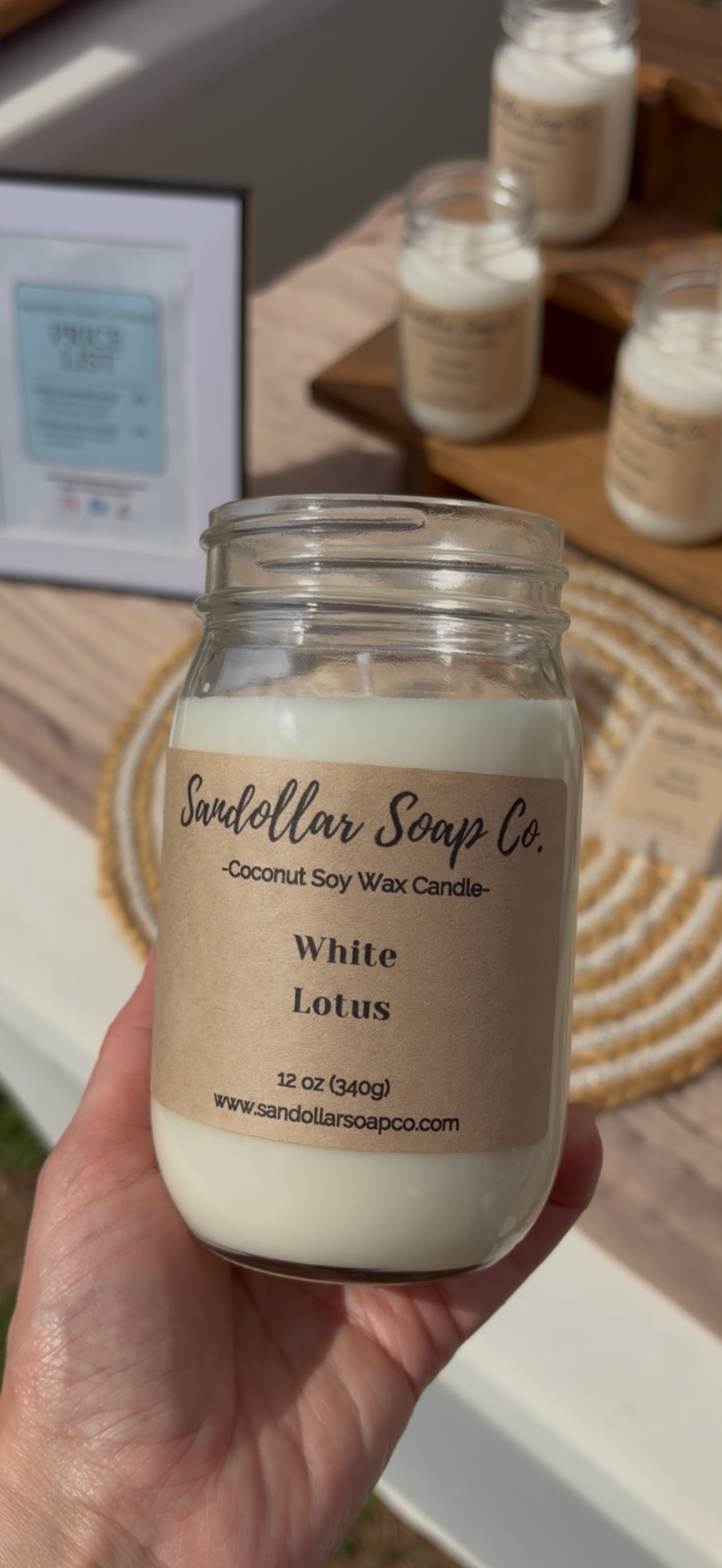 Hand holding a candle labeled 'White Lotus' from 'Sundellar Soap Co.' with more candles in the background.